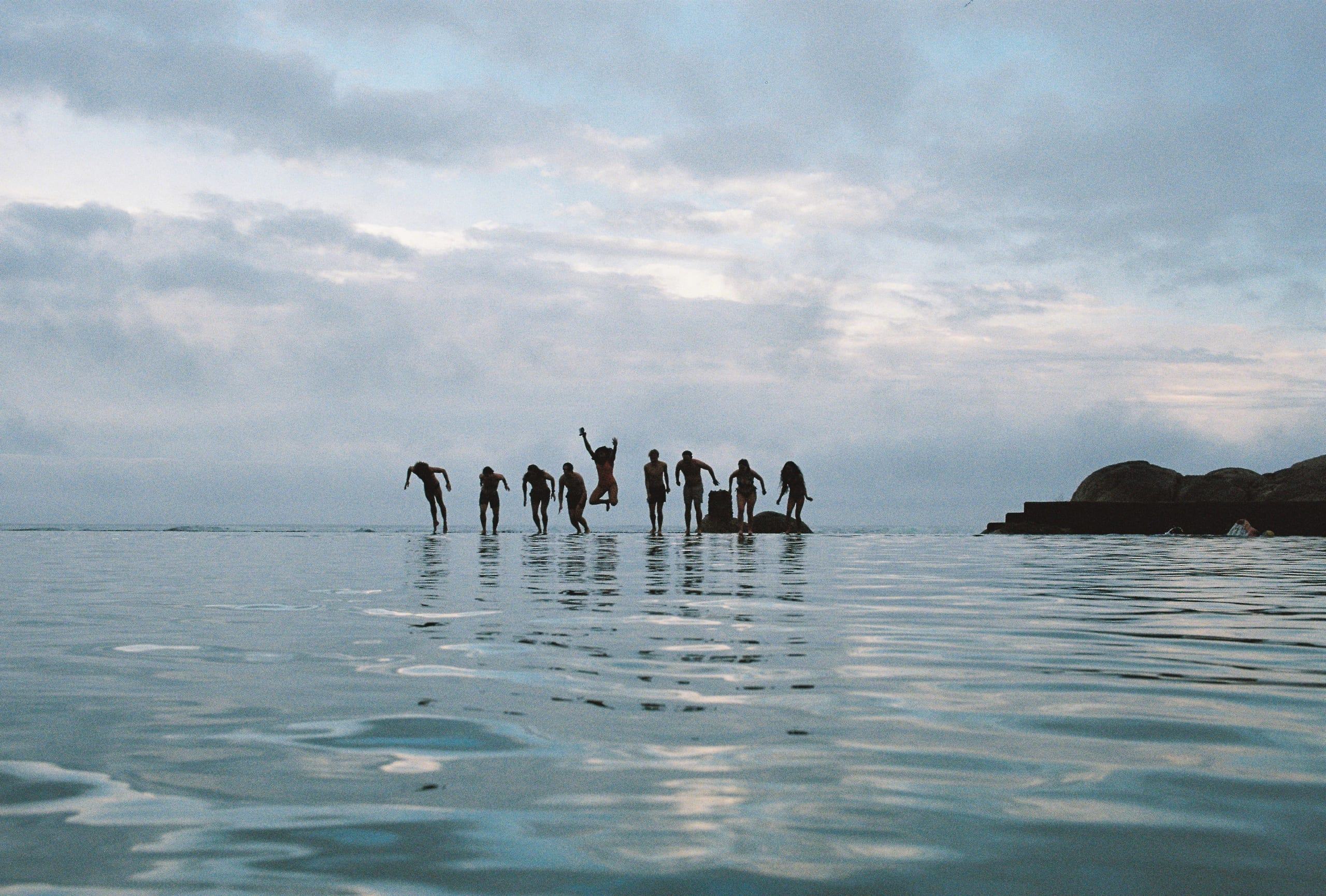 A group of swimmers jumping into the ocean from a rock, photographed from the water.